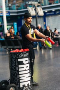 A man presents Prime beverages indoors, engaging the audience at a lively promotional event.